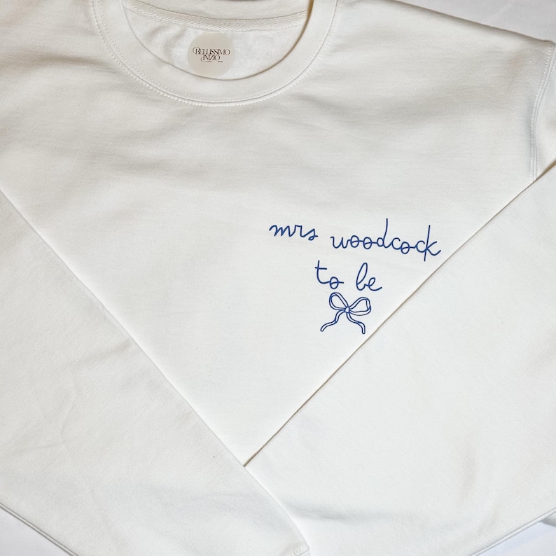 May include: A white crew neck sweatshirt with the text "mrs woodcock to be" in navy blue script, along with a bow graphic. The sweatshirt has a small label near the neckline.