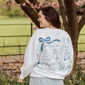 May include: White sweatshirt with a blue ribbon and the phrase "going to the chapel" with wedding-themed illustrations. The design includes a cake, champagne flutes, and a car. The person is wearing striped pants.