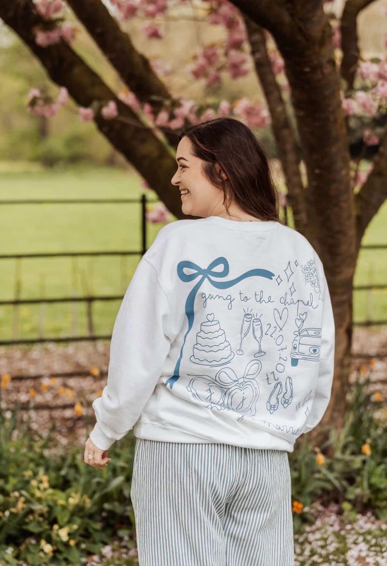 May include: White sweatshirt with a blue ribbon and the phrase "going to the chapel" with wedding-themed illustrations. The design includes a cake, champagne flutes, and a car. The person is wearing striped pants.