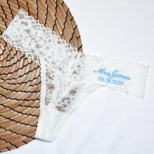 May include: White lace thong underwear with scalloped edges. The text "Mrs. Jones" and the date "06.08.2026" are embroidered in light blue. The underwear is displayed on a woven, circular mat.
