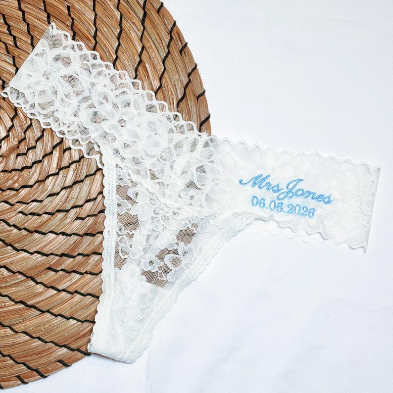 May include: White lace thong underwear with scalloped edges. The text "Mrs. Jones" and the date "06.08.2026" are embroidered in light blue. The underwear is displayed on a woven, circular mat.