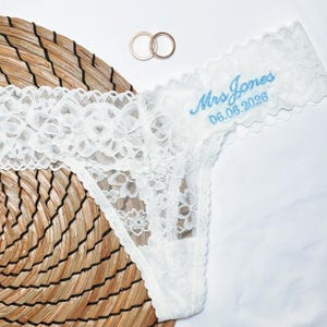 May include: White lace thong underwear with the embroidered text "Mrs Jones 06.06.2026" in light blue. Two gold wedding rings are placed above the underwear. The underwear is on a white surface and a woven mat.