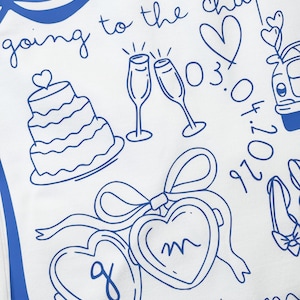May include: White fabric with blue line art featuring wedding-themed illustrations. Includes a tiered cake, champagne flutes, hearts, and the phrase "going to the chapel." Also includes the date 03.04.2026 and initials in heart-shaped lockets.