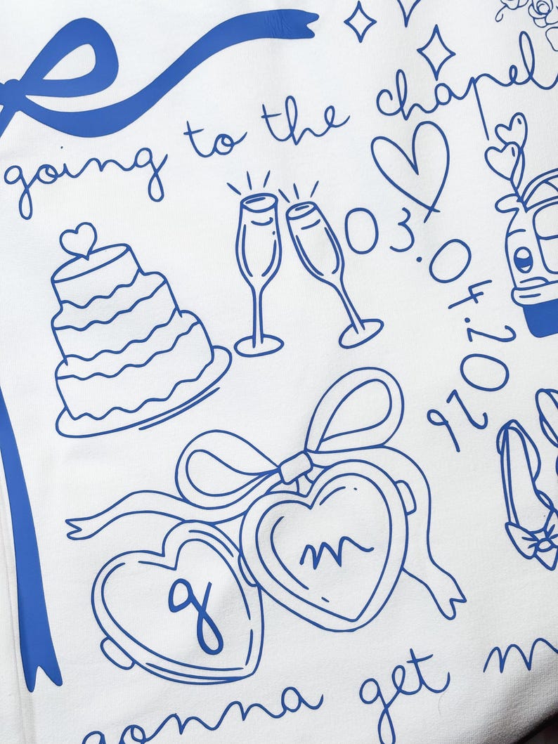 May include: White fabric with blue line art featuring wedding-themed illustrations. Includes a tiered cake, champagne flutes, hearts, and the phrase "going to the chapel." Also includes the date 03.04.2026 and initials in heart-shaped lockets.
