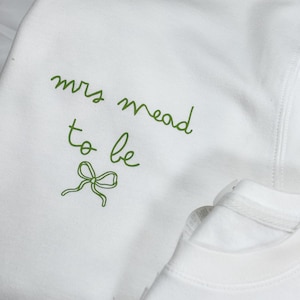 May include: White sweatshirt with green cursive text that reads "mrs mead to be" and a green bow graphic. The sweatshirt is made of a soft material and is likely for a bridal event.