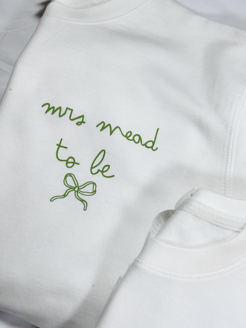 May include: White sweatshirt with green cursive text that reads "mrs mead to be" and a green bow graphic. The sweatshirt is made of a soft material and is likely for a bridal event.