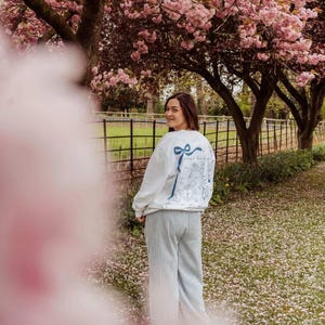 May include: A person wearing a white sweatshirt with a blue bow and text design on the back, paired with striped pants. The scene is set outdoors under pink flowering trees, with a fence and grass in the background.