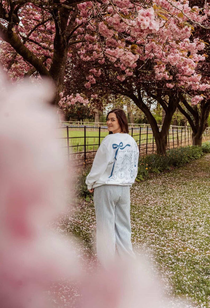 May include: A person wearing a white sweatshirt with a blue bow and text design on the back, paired with striped pants. The scene is set outdoors under pink flowering trees, with a fence and grass in the background.