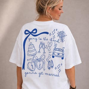 May include: White t-shirt with a blue graphic design featuring wedding-themed illustrations and text. The design includes a cake, champagne glasses, a car, and the words "going to the chapel" and "gonna get married".
