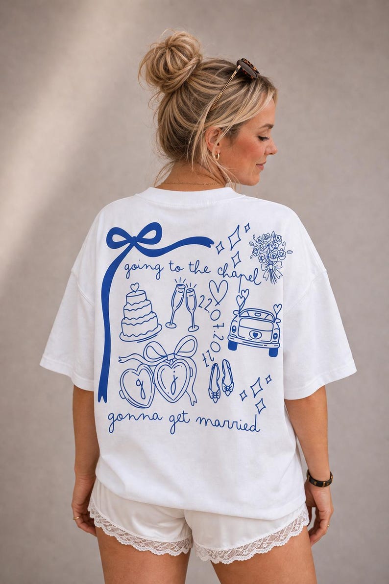 May include: White t-shirt with a blue graphic design featuring wedding-themed illustrations and text. The design includes a cake, champagne glasses, a car, and the words "going to the chapel" and "gonna get married".