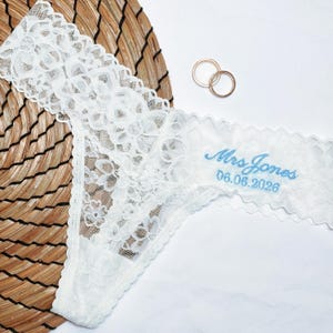 May include: White lace thong underwear with the embroidered text "Mrs Jones 06.06.2026" in light blue. Two gold rings are in the background. The underwear is on a white surface with a woven mat.