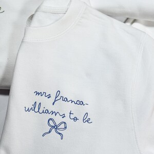 May include: White crew neck sweatshirt with the text "mrs franca-williams to be" in blue script, accompanied by a blue bow illustration. Another sweatshirt is partially visible in the background.