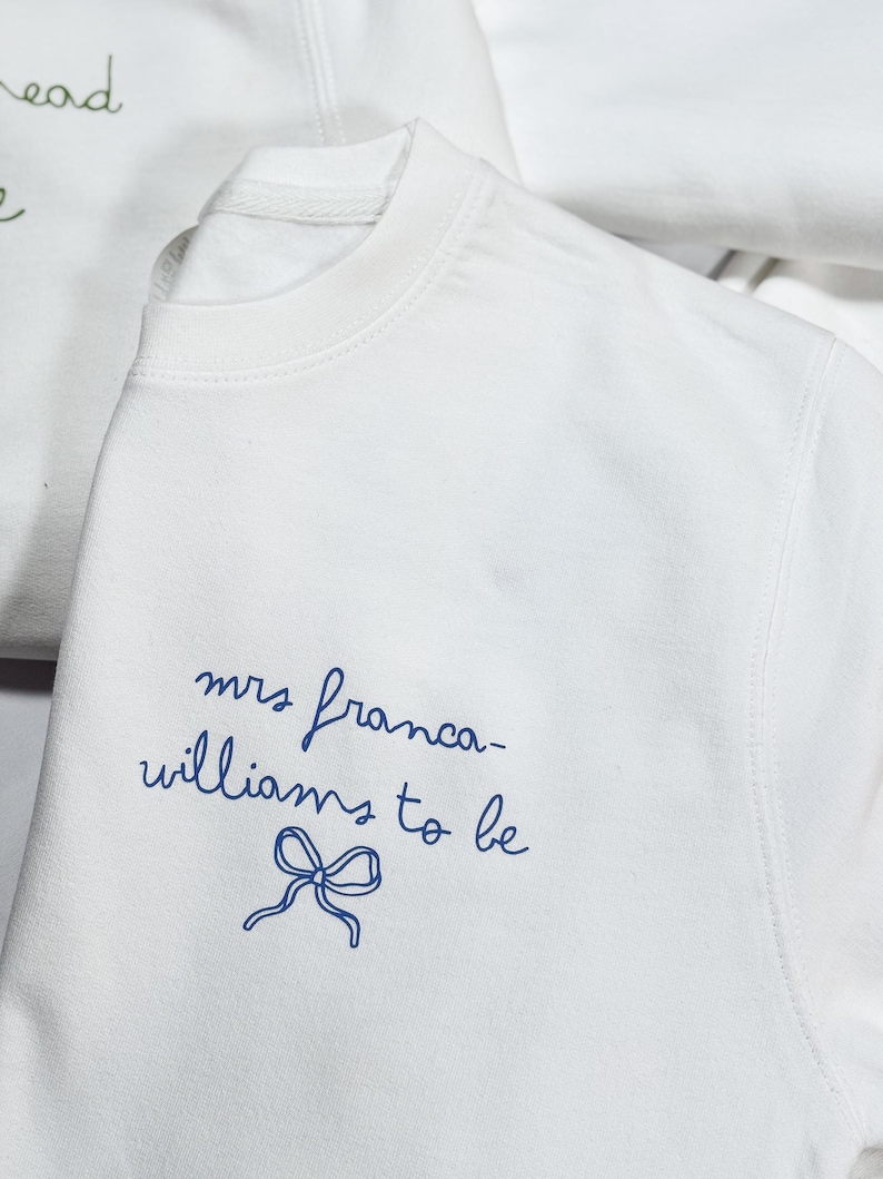 May include: White crew neck sweatshirt with the text "mrs franca-williams to be" in blue script, accompanied by a blue bow illustration. Another sweatshirt is partially visible in the background.