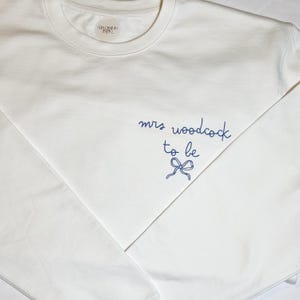 May include: A white crew neck sweatshirt with the text "mrs woodcock to be" in navy blue script, along with a bow graphic. The sweatshirt has a simple, clean design, perfect for a bridal event.