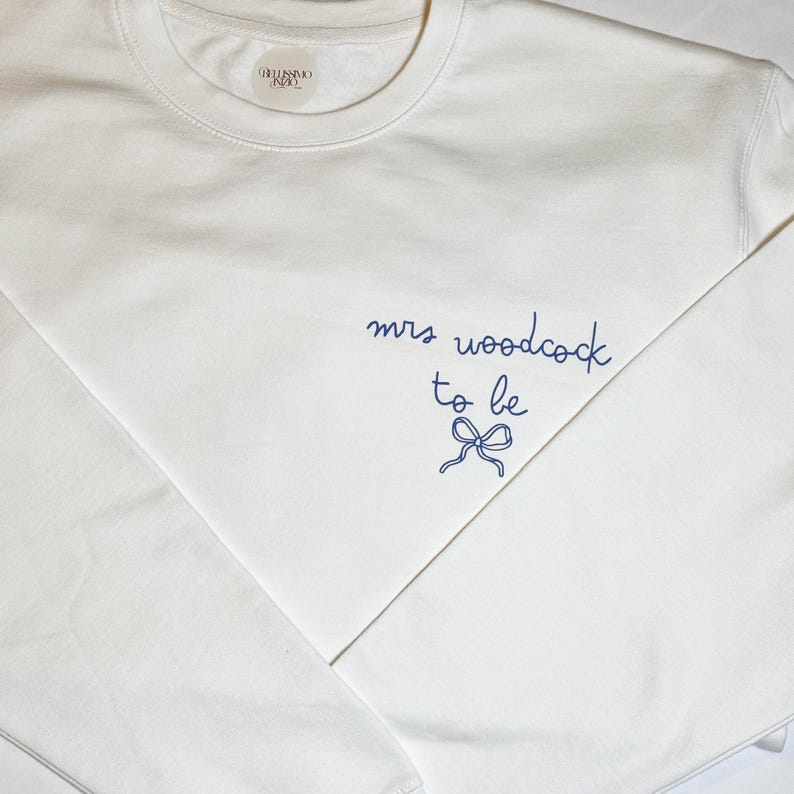 May include: A white crew neck sweatshirt with the text "mrs woodcock to be" in navy blue script, along with a bow graphic. The sweatshirt has a simple, clean design, perfect for a bridal event.