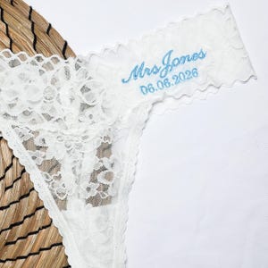 May include: White lace thong underwear with a delicate floral pattern. The text "Mrs Jones 06.06.2026" is embroidered in light blue on the waistband. The underwear is displayed on a woven surface.