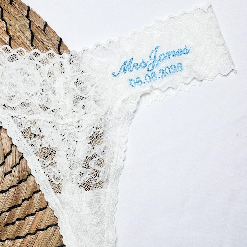 May include: White lace thong underwear with a delicate floral pattern. The text "Mrs Jones 06.06.2026" is embroidered in light blue on the waistband. The underwear is displayed on a woven surface.