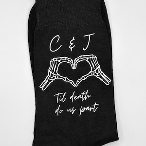 May include: Black socks featuring a white skeleton hand heart design and the text "C & J" above "Til death do us part". The socks are made of a dark material, suitable for everyday wear.
