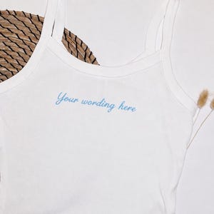 May include: A white vest top with thin shoulder straps featuring the text Your wording here embroidered in light blue cursive script across the chest. The garment is made of a plain white fabric and displays a clean, simple design.