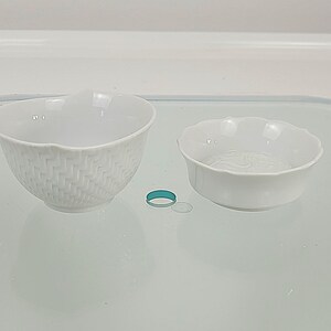 May include: Two white porcelain bowls. One bowl has a woven texture, and the other has a scalloped edge. A small teal ring and a clear plastic disc are between the bowls. The bowls are on a reflective surface.