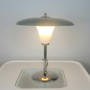 Puede incluir: Una lámpara de mesa moderna con base y tallo plateados, coronada por una pantalla verde claro en forma de platillo. La lámpara presenta un difusor de luz de vidrio esmerilado en forma de tulipán y está iluminada, emitiendo un brillo cálido.