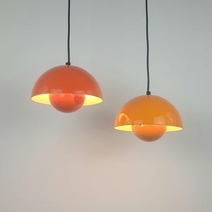 Peut inclure: Deux lampes suspendues orange au design rétro. Chaque lampe a un abat-jour arrondi en forme de dôme et une ampoule sphérique. Les lampes sont suspendues à des cordons noirs sur un fond neutre. L'intérieur des abat-jours est d'une couleur plus claire.