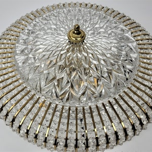 Op de afbeelding: Een ronde, goudkleurige plafondlamp met een kristalontwerp. De lamp heeft een centrale koepel met een kristalpatroon en een ring van kristallen rond de rand.