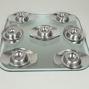 Op de afbeelding: Een set van zeven zilverkleurige eierdopjes, elk met een afgeronde kom en een ovale basis. De eierdopjes staan op een heldere glazen tray, die het licht reflecteert. Het ontwerp is eenvoudig en elegant.