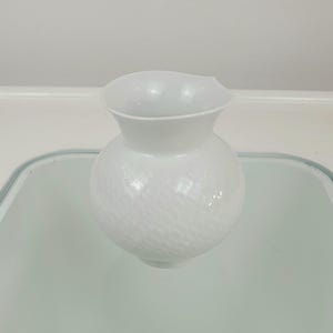 May include: A white, textured glass vase with a round body and a flared, slightly asymmetrical rim. The vase has a detailed, woven-like pattern across its surface, adding visual interest to the simple, elegant design. The vase is likely used for decorative purposes.