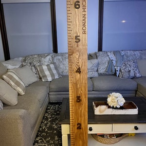May include: A wooden growth chart with engraved height markings from 1 to 6 feet. The chart is made of light-colored wood and features the name "RONAN MICHAEL" vertically. The numbers are dark brown, and the design is simple and elegant.