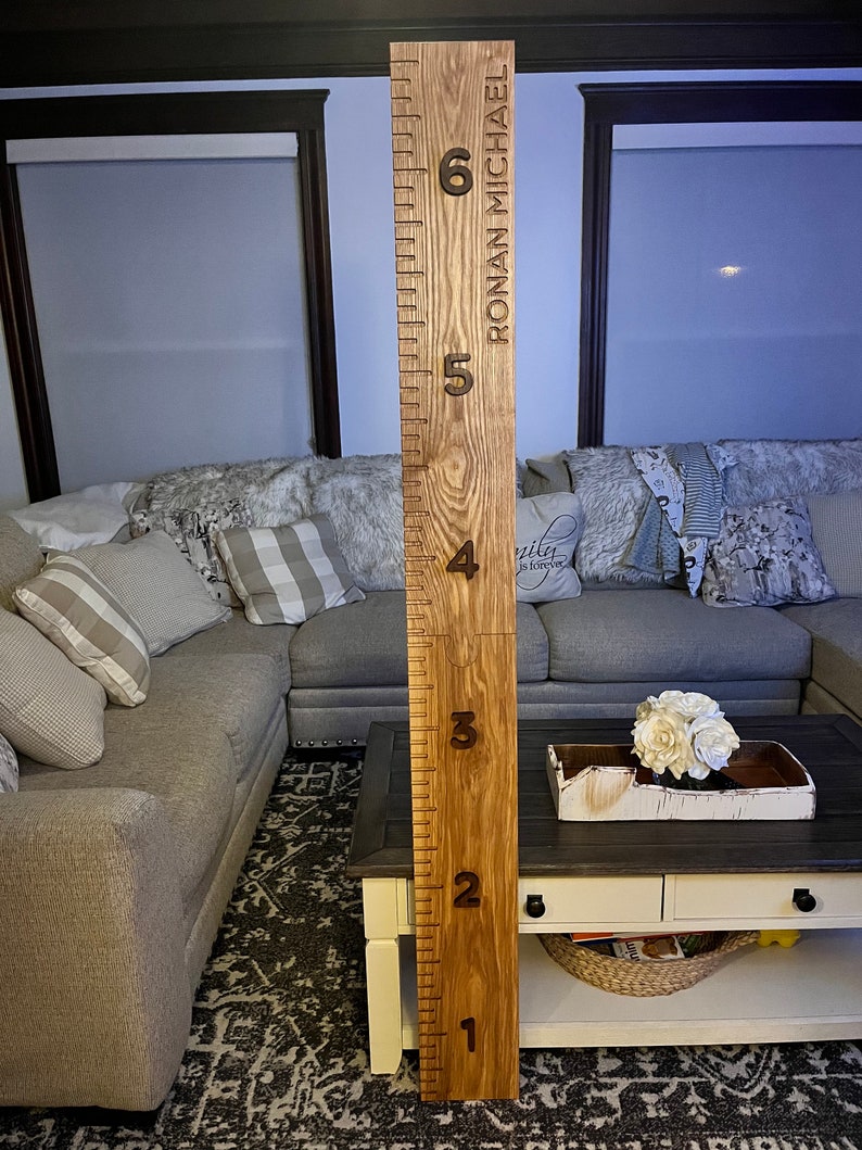 May include: A wooden growth chart with engraved height markings from 1 to 6 feet. The chart is made of light-colored wood and features the name "RONAN MICHAEL" vertically. The numbers are dark brown, and the design is simple and elegant.