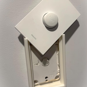 May include: A white Philips Hue dimmer switch, partially removed from its wall mount. The switch has a circular button with the word "hue" and the Philips logo. The wall mount is also white.