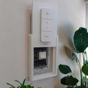 May include: A white Philips Hue smart light switch with a white remote control. The remote control has a power button, a brightness control, and a button for colour temperature. The switch is mounted on a white wall.