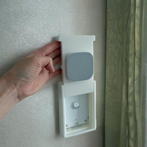 May include: A white smart home device with a grey control panel is mounted on a wall. The device has a rectangular base and a rounded top with two buttons and a circular button. The device is designed for home automation.