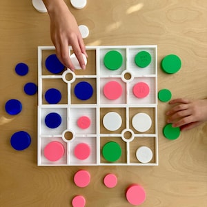 May include: A white board game with a grid of squares, filled with blue, green, pink, and white circular game pieces. Some pieces have the words "YOU LOSE" printed on them. Hands are placing the game pieces on the board.