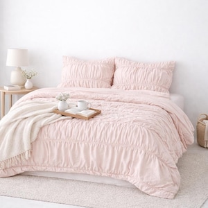 May include: A light pink bedspread and pillow set with a textured, ruched design. A cream-colored throw blanket is draped over the bed. A wooden tray with two mugs and a book sits on the bed. A small side table with a lamp and vase is next to the bed.