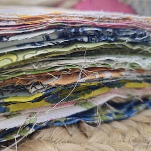 May include: A close-up shot of a pile of patterned fabric remnants. The fabrics feature a range of colours and designs, including floral and abstract patterns. The edges of the fabric are frayed, and the stack rests on a textured surface.