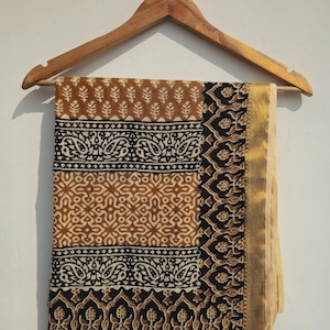 Könnte beinhalten: Ein gemustertes Textil in Braun und Schwarz, das an einem Holzbügel hängt. Das Design umfasst Blatt- und Paisley-Motive in Braun, Schwarz und Weiß. Ein goldfarbener Rand säumt den Stoff.