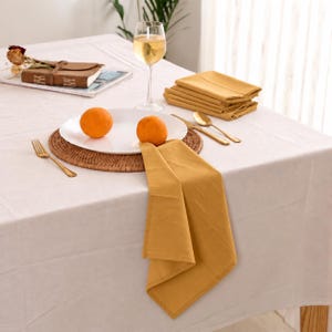 Könnte beinhalten: Ein Set aus senfgelben Stoffservietten, ein weißer Teller mit zwei Orangen, eine goldene Gabel, ein goldener Löffel und ein goldenes Messer. Der Tisch ist mit einer weißen Tischdecke bedeckt und ein Weinglas mit heller Flüssigkeit steht daneben.