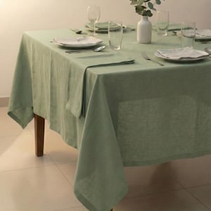 Op de afbeelding: Een saliegroene linnen tafelkleed bedekt een houten tafel die gedekt is voor een maaltijd. De tafel is gedekt met witte borden, bestek, heldere drinkglazen en een kleine witte vaas met groen. Het tafelkleed is ongeveer 183 cm vierkant.