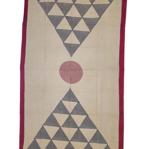 Puede incluir: Una manta de tejido color crema con un borde rojo. La manta presenta un diseño geométrico de triángulos negros y blancos y un círculo rojo en el centro.