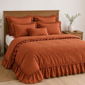May include: A rust-coloured bedding set featuring a duvet cover and multiple pillows, all adorned with ruffled edges. The set includes two large square pillows, two rectangular pillows, and one smaller lumbar pillow, all in a solid burnt orange hue.