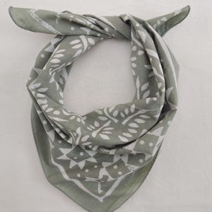 Könnte beinhalten: Salbeigrünes Bandana mit weißem Blatt- und geometrischem Muster. Das quadratische Tuch ist gefaltet und gebunden, wodurch der detaillierte Druck zur Geltung kommt. Der Stoff wirkt weich und leicht, geeignet für verschiedene Zwecke.