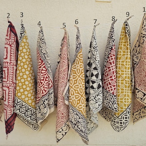 Op de afbeelding: Een verzameling van elf katoenen bandanas met patronen die tegen een witte muur hangen. De bandanas hebben verschillende ontwerpen in kleuren als zwart, rood, geel en wit. Elke bandana is ongeveer 53 cm vierkant.