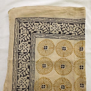Puede incluir: Textil beige plegado con un borde floral negro y blanco y un patrón repetido de rayos de sol amarillos con centros negros. El textil es probablemente una bufanda o un paño decorativo.
