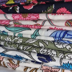 Może przedstawiać: Stos tkanin we wzory kwiatowe. Tkaniny prezentują różowe, niebieskie, zielone i żółte wzory kwiatowe na białym tle. Wzory kwiatowe różnią się rozmiarem i stylem, tworząc wizualnie zróżnicowaną kolekcję.