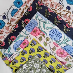 Może przedstawiać: Kolekcja złożonych kwadratów materiału z kwiatowymi wzorami w różnych kolorach. Wzory obejmują niebieskie, różowe i żółte kwiaty na białym, granatowym i zielonym tle. Tkaniny ułożone są w warstwową, ukośną kompozycję.