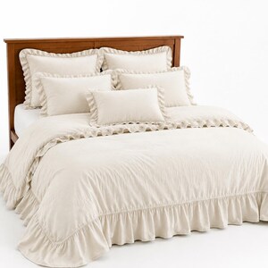 Pode incluir: Um conjunto de roupa de cama na cor creme, composto por uma capa de edredão e cinco almofadas, todas decoradas com bordas de folhos. O conjunto está exposto numa estrutura de cama de madeira, exibindo um acabamento em tecido texturado.