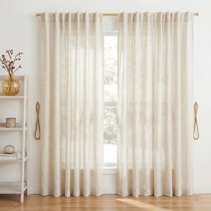 Könnte beinhalten: Beige Leinen-Vorhänge, die an einer goldenen Stange hängen und ein Fenster einrahmen. Die Vorhänge werden mit Kordel-Raffhaltern zurückgehalten. Ein weißes Bücherregal mit Dekorationsartikeln ist sichtbar.