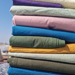 Könnte beinhalten: Ein Stapel bunter Stoffrollen in verschiedenen Farbtönen, darunter Blau, Grün, Gelb, Rosa und Lila. Der Stoff scheint eine Naturfaser zu sein, möglicherweise Leinen oder Baumwolle, mit einer leicht strukturierten Oberfläche. Die Rollen sind vor einem blauen Himmel gestapelt.